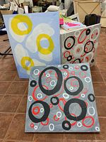 Three abstract paintings on floor with Baltic Birch panel visible, showing colors including yellow, gray, black, red, and white.