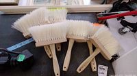 Six large white synthetic bristle brushes with wooden handles, some bristles tipped in orange, laid out on a surface.
