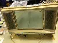 Full view of the vintage cosmetic magnifying mirror, showing the rectangular magnifying mirror between two decorative gold-tone side lights with a beige plastic frame and base.