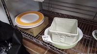 Photo showing the yellow rimmed Villeroy & Boch plates stacked on a box and the square white ceramic plant pot placed on the pale green Pyrex baking dish, all resting on metal shelving.