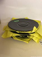 Six black glass round plates stacked with yellow packing paper between them, showing the smooth glossy finish and round shape.