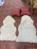 Two white sheep skin rugs side-by-side with two red pillows behind them on tiled floor.