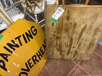 Photo showing yellow painted wooden sign with black text 'PAINTING OVERHEAD' with visible paint stains