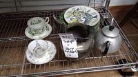 Teacups, collector plates, aluminum teapot, silver plate pitcher, and metal tea strainer arranged on metal rack.