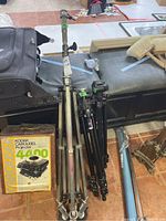 Three tripods and film light stand arranged on floor. Faniro Quartz Color Italia tripod on wheels to the left, two smaller tripods (Gruppi Manfoto and Bausch & Lomb) to the right, Kodak Carousel projector box visible but projector not included.