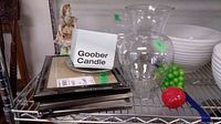Shelf holding two clear glass vases, glass fruit including green grapes and a red apple, three stacked black picture frames, a white container labeled 'Goober Candle', and a vintage figurine with visible damage.