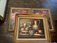 Three framed art pieces laid on floor, two identical floral prints and one still life painting with fruit and a jug.