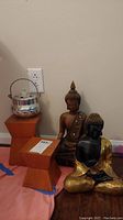 Two Buddha figures seated on orange wooden pedestals next to a silver tin pot on the floor against the wall