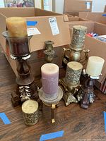 Seven candle holders and six candles arranged on wooden floor with cardboard boxes in background