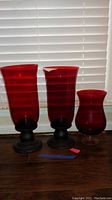 Three red glass candle holders of varying heights placed on wood floor in front of white blinds, two with black bases and one with clear base.