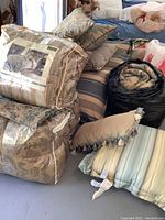 Photo showing four queen comforters stacked including some in plastic packaging, and several matching decorative throw pillows, all placed on and around a bed with a striped mattress cover.