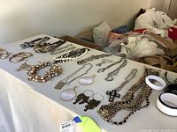 Image showing multiple necklaces, bracelets, and two pairs of earrings arranged on a white surface. Jewelry includes gold-tone and silver-tone metal chains, beaded pieces, and earrings with coin dangles and cross pendants.