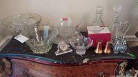 A collection of crystal and glass bowls, decanters, salt and pepper shakers on a wooden dresser including boxed Mikasa Celebrations bowl, clear glassware with etched leaf pattern, Coppercraft Guild metal shakers, and a small decorative clock.