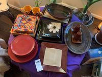 Varied dinner plates and servingware including square and round plates in multiple colors and patterns, mugs, serving platters, and salt and pepper shakers arranged on table.