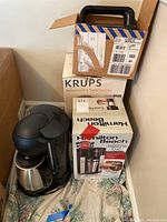 Four coffee makers stacked including boxed Hamilton Beach and Krups boxes with two black Krups coffee makers with stainless steel thermal carafes visible.