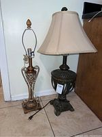 Two table lamps placed side by side on a tiled floor, one with dark ornate base and cream shade, the other with copper and gold metal base without shade.