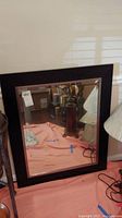 Square wall hanging mirror placed upright on the floor against a wall, showing reflections and some surrounding objects including lamp and table. Visible scratches on frame.