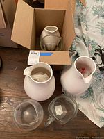 Six pitchers including three glass and three ceramic placed on a wooden surface with one ceramic pitcher wrapped and boxed.