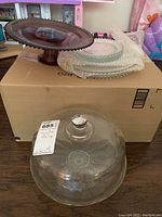 Purple hobnail glass cake stand on left, stacked clear hobnail glass serving plates on right, and a clear glass dome lid on top of a box.