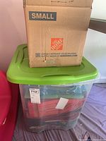 Green lidded plastic tote with a cardboard box on top, containing linens inside. Tote is labeled with an auction tag.