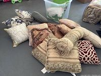 Sixteen assorted throw pillows piled on floor, showing varied colors and designs, also glimpse of a large green plastic tote.