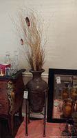 Full view of tall brown crackled vase with dried grass inside. Vase is held within four-legged metal stand.