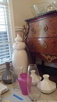 Seven vases grouped together showing variety of shapes and colors, placed on floor beside furniture near window with natural light