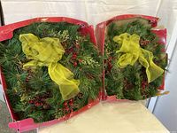 Two large Christmas wreaths in red boxes, showing green pine needles, pinecones, red berries, and yellow decorative bows.