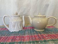 Two teapots placed side by side on a multicolored woven surface, showing their shapes, handles, spouts and lids.