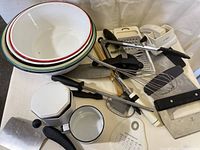 Four white nesting graniteware bowls stacked with various kitchen utensils underneath and around them, showing wear and some dirt.