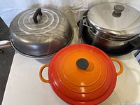 Top view showing Le Creuset orange enameled cast iron pot with lid, stainless steel pot with lid, and large metal pan with handle