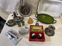Metal wind chimes with green pan, white enamel roasting pan, decorative metal ashtray, pair of Yin and Yang balls with red box, metal candle holder, metal condiment set, metal clamp tool, and wooden utensil displayed on table.