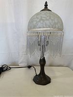 Full view of the table lamp showing white frosted glass shade with beaded fringe and bronze-tone base.