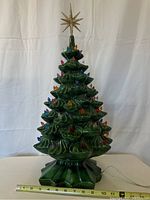 Front view of the green ceramic Christmas tree on its base with plastic light inserts and gold glitter star topper. Measuring tape shows base width.