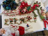 Full view of various vintage Christmas items including elf clips, Santa sleigh, wreath, and dolls laid out on table.