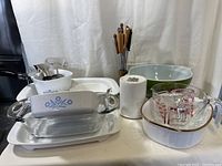 Overview of kitchenware lot including Corning Ware baking dishes with blue cornflower pattern, Pyrex measuring cups, green Corning Ware bowl, and Portmeirion botanical storage container with lid, along with metal skewers with wooden handles.
