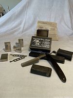 Wide view of entire shaving kit showing straight razor, metal blade cases, sharpening block, Stroppe Razor Sharpener box with instructions, and other metallic accessories