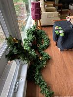 Photo showing three artificial evergreen garlands resting by windowsill and floor. Green with gold flower accents, approx 9 feet long each, slightly dusty.