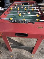 Foosball table showing front side, wooden body painted red, scoring sliders, plastic players in yellow and red uniforms on metal rods.