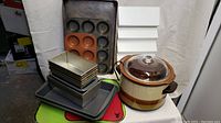 Wide angle photo showing ceramic slow cooker, multiple metal loaf pans, two muffin pans, and two large baking sheets stacked and arranged.