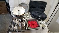 Wide view of kitchen utensils tray with silverware, multiple baking pans, red plastic cookie cutters, and covered roasting pan on table.