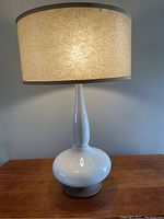 Photo of the full lamp turned on showing white ceramic base with slender neck and cream colored shade that is lit causing the crazing and discoloration of the shade to be visible.