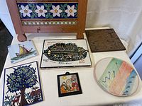 Overview of tiles and trivets laid out on fabric background. Shows various decorative tiles including map tile, tree tile, ship tile, religious icon tile, round abstract trivet, and brown mottled trivet, along with wood-framed floral tile wall hanging in background.