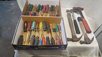 Box with approximately 32 screwdrivers of various sizes and handle colors plus three hammers and one metal pry bar arranged side by side on a white cloth.