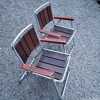 Pair of vintage folding chairs showing wooden slat seats and backs with reddish wooden armrests on a silver metal frame, placed outdoors on gravel.