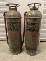 Front view of two copper-bodied, vintage Pyrene soda-acid fire extinguishers standing side by side, showing embossed branding and attached hoses.