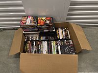 Wide top view of large cardboard box filled with DVDs and Blu-Ray movies stacked horizontally and vertically. Covers and titles visible on discs.