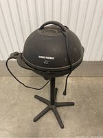 Front view of George Foreman electric BBQ grill on stand with cord wrapped around it.