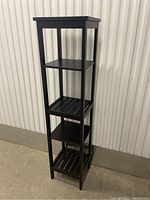 Tall black shelving unit with solid and slatted shelves against a corrugated metal wall.