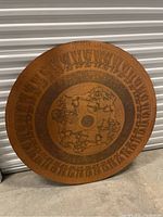 Full view of large circular copper art piece with patterned border and central figures in wheel contraptions.